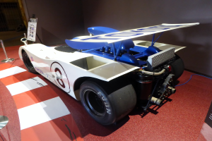 Ein weiß-blauer Rennwagen steht in einem Museum ausgestellt, umgeben von Absperrbändern und Schautafeln, mit einer Wand im Hintergrund, die auf das Goodwood Festival of Speed hinweist.