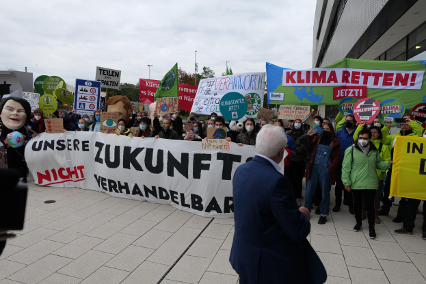 Eine Gruppe von Menschen mit Schildern und Transparenten protestiert gegen den Klimawandel vor einem Gebäude in Deutschland, mit einer Person, die links eine Kamera hält, Bäumen, Pfählen und einem klaren blauen Himmel im Hintergrund.
