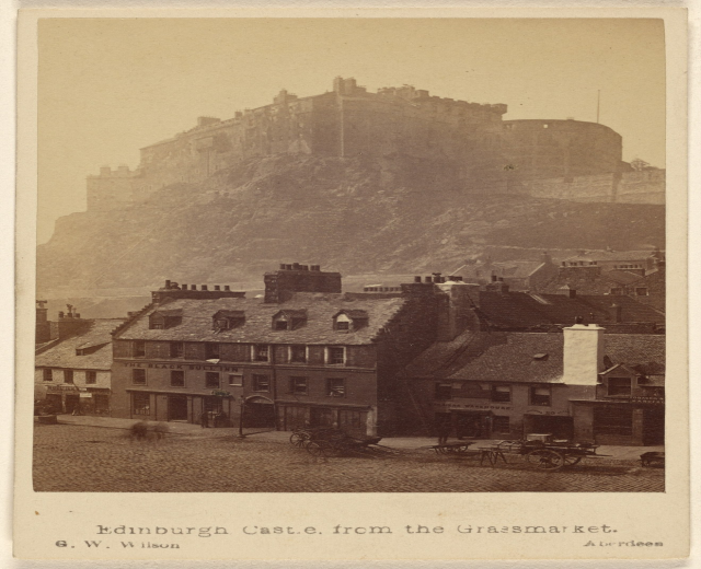 Alte Fotografie des Edinburgh Castle von Grassmarket aus betrachtet, mit Gebäuden und Karren im Vordergrund und einem Hügel im Hintergrund; Text unten.