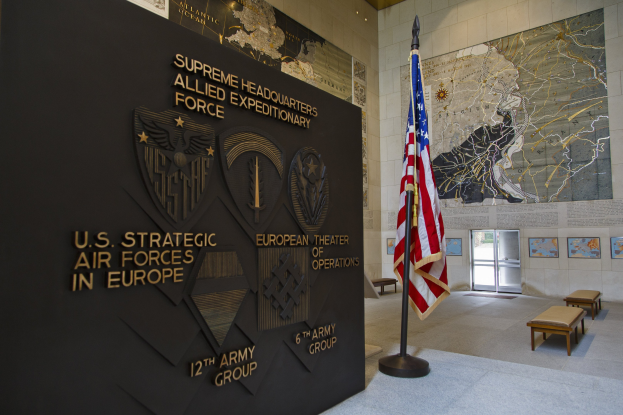 Eintritt zum U.S. Strategic Air Forces Headquarters in Europa, mit einer amerikanischen Flagge, einer Wand mit Text und Logos, Bänken, einer Tafel, Fotorahmen, beleuchteter Decke und einer Tür im Hintergrund.