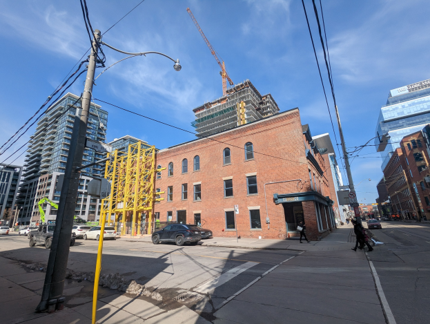 Eine belebte Stadtstraße mit fahrenden Fahrzeugen, Fußgängern auf dem Gehweg, Stromleitungen, Straßenlaternen und Gebäuden, mit einem entfernten Baugerüst mit einem Kran gegen einen bewölkten Himmel.