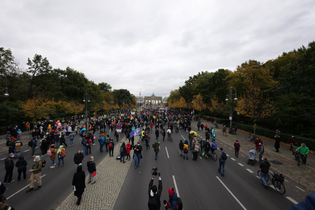 Eine große Gruppe von Menschen marschiert eine baumbestandene Straße in Berlin entlang, mit Kameras bewaffnet, vor einem Gebäude und einem klaren Himmel.