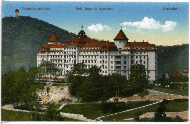 Eine alte Postkarte zeigt das Hotel Imperial in Karlsbad, Deutschland, ein großes Gebäude mit zahlreichen Fenstern, umgeben von Bäumen, Pflanzen und Gras, mit einem Weg im Vordergrund, Hügeln im Hintergrund und einem bewölkten Himmel, mit Text oben auf dem Bild.