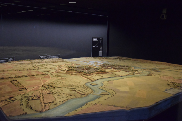 Ein Stadtmodell in einem Museum mit einem Tisch im Vordergrund und einer Texttafel im Hintergrund, unter warmen Deckenleuchten.