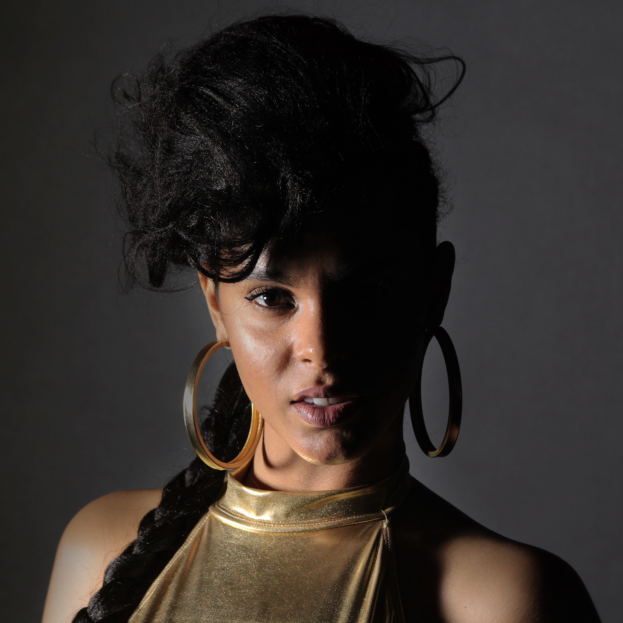 Eine Frau in einem goldenen Kleid und großen Ohrringen steht selbstbewusst vor einer Wand, ihre Haare sind zu einem sleeken Bob gestylt, sie trägt dezentes, natürliches Make-up mit pinkem Rouge und glänzenden Lippen, sieht direkt in die Kamera und hat einen entschlossenen Gesichtsausdruck.