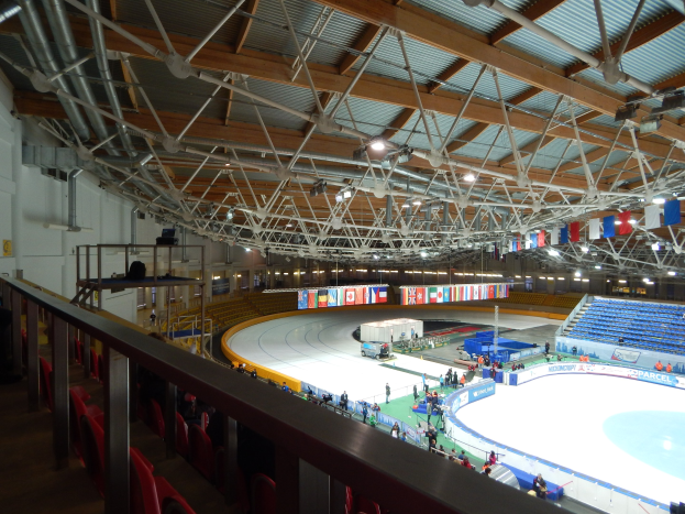 Großes Indoor-Eisstadion mit zahlreichen Menschen auf dem Eis, umgeben von Sitzplätzen, Geländern, Brettern, Leuchten und strukturellen Elementen, mit einer Wand im Hintergrund.