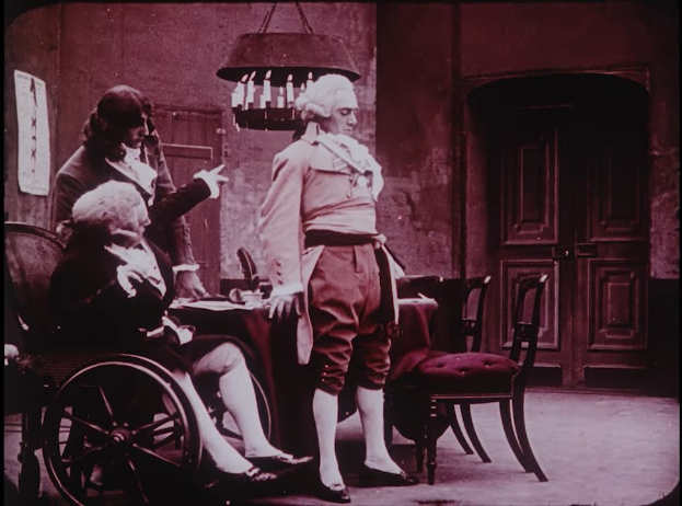 Ein altes Foto eines Mannes im Rollstuhl, der mit zwei Frauen spricht, die um einen Schreibtisch mit verschiedenen Gegenständen sitzen, mit einem Stuhl daneben, einer Tür und einem Kronleuchter im Hintergrund.