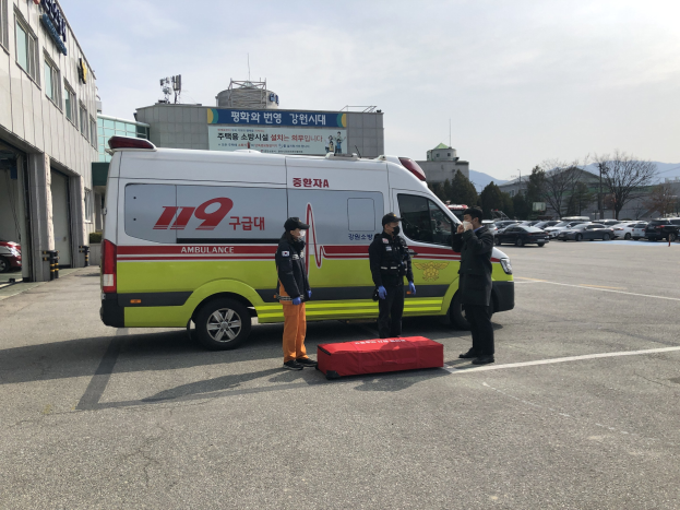 Ambulanz auf einem Parkplatz neben einem Gebäude mit drei Personen davor geparkt, links ein Gebäude mit Glasfenstern, andere Fahrzeuge, Bäume, Berge und Himmel im Hintergrund.