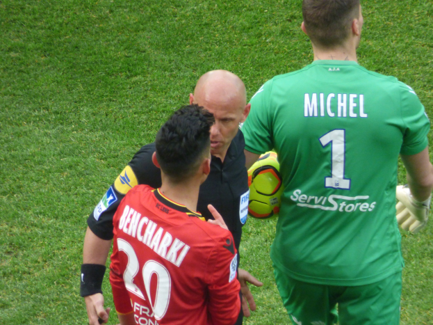 Schiedsrichter spricht mit zwei Fußballspielern auf einem grünen Rasen, ein Spieler hält einen Ball.