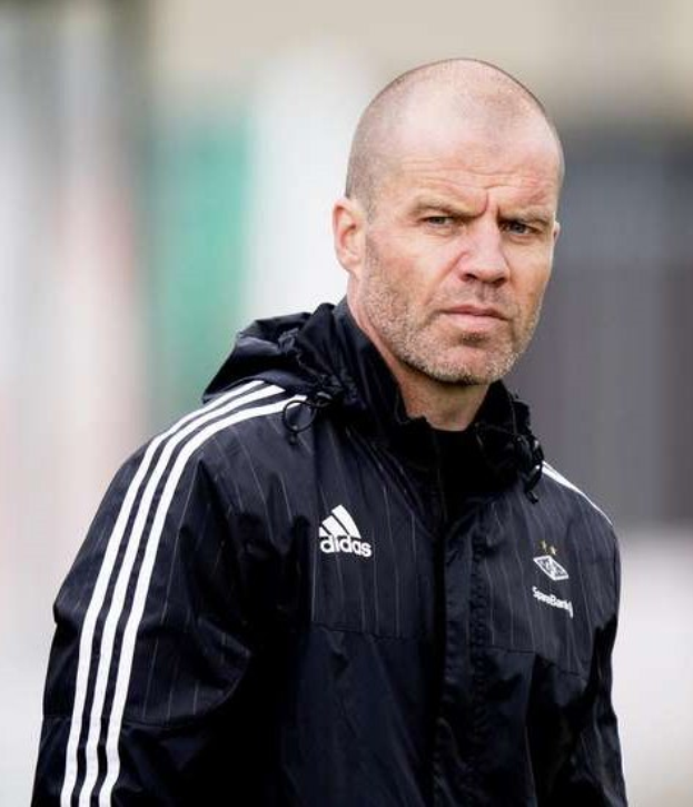 Glatzköpfiger Mann in einem blauen Jackett mit weißen Streifen und weißem Kragen, der auf einem Fussballfeld steht und die Hände verschränkt, ernsthafter Ausdruck, unscharfer Hintergrund.