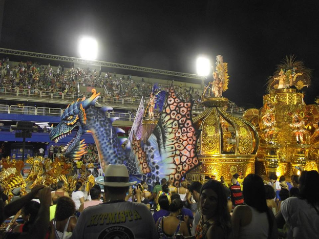 Eine große Menschenmenge versammelt sich um ein hell erleuchtetes, buntes Floß auf einem Volksfest, mit einem Geländer und einem Gebäude im Hintergrund unter einem dunklen Himmel.