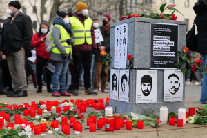 Eine Gruppe von Menschen steht feierlich vor einem mit Kerzen, Blumen und Plakaten geschmückten Denkmal, umgeben von Bäumen und Gebäuden mit Fenstern im Hintergrund, und gedenkt eines Dramas in Berlin, Deutschland.