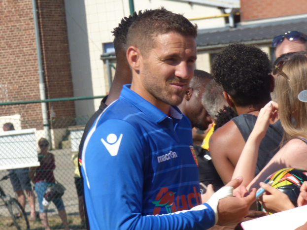 Ein Mann in einem blauen Hemd signiert Autogramme für eine Gruppe von Menschen, einige tragen Mützen und Schutzbrillen, mit einem Gebäude, einem Maschendrahtzaun und einem Fahrrad im Hintergrund.