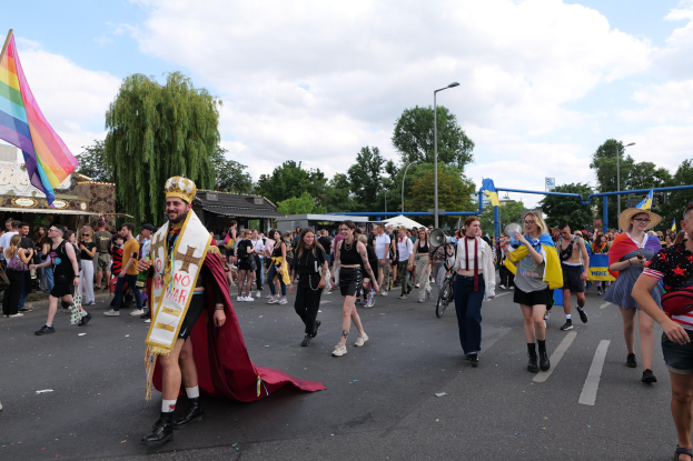 Eine Gruppe von Menschen marschiert bei der Gay Pride Parade 2018 mit einer Regenbogenfahne und Musikinstrumenten, im Hintergrund sind Laternenpfähle, Bäume, Schuppen und ein bewölkter Himmel zu sehen.