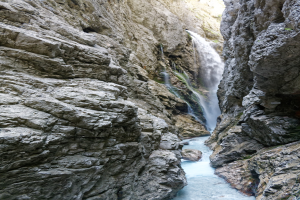 Kleiner Wasserfall, der an zerklüfteten Felsen in einem steinigen Tal herabfließt, umgeben von grünen Hügeln unter strahlendem Sonnenlicht.