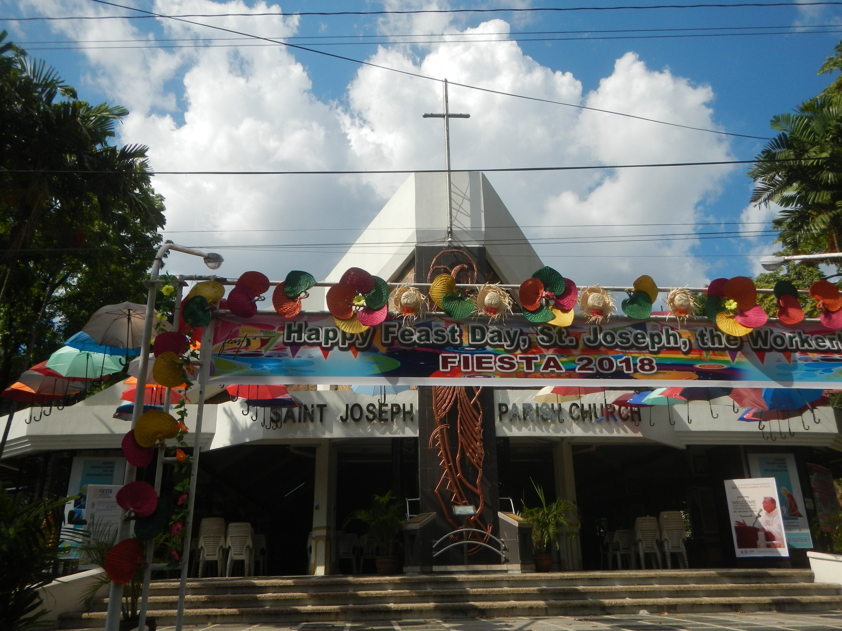 Ein Gebäude mit einem Schild, das "Frohes Fest St. Josef der Arbeiter" trägt, davor stehend, mit einer Treppe zum Eingang, einem Bogen mit hängenden Dekorationen, Außenbestuhlung, Topfpflanzen, Bäumen und einem bewölkten Himmel.