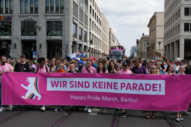 Eine Gruppe von Menschen marschiert mit einem pinken 'Happy Pride March'-Schild die Straße in Berlin entlang, mit Gebäuden, Laternenmasten und Verkehrszeichen an der Straße unter einem bewölkten Himmel.