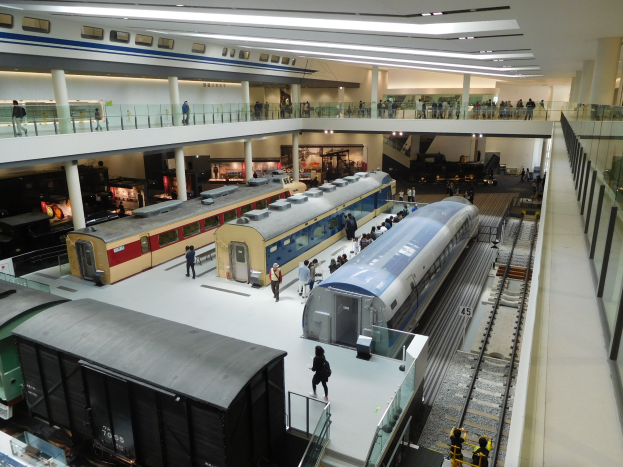 Eine große Museumsausstellung zeigt verschiedene Züge in unterschiedlichen Größen und Farben, bei der Besucher vom Boden, Geländern und Säulen unter Deckenbeleuchtung zuschauen.