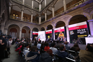 Eine Gruppe von Menschen, die an einem Tisch mit einem Podium und einem Mikrofon vor einer Menge, Kameras und Bannern sitzen, was eine Pressekonferenz für das CDMX 2017-Event vermuten lässt.