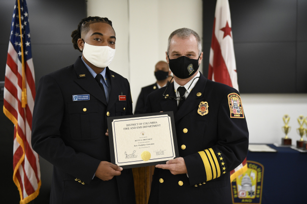 Zwei Männer in Feuerwehranzügen, einer identifiziert als Leiter der Feuerwehr des District of Columbia und der andere als Stellvertreter, stehen nebeneinander und tragen Masken und halten ein Zertifikat, mit einem Tisch voller Auszeichnungen, Schilde und Flaggen im Hintergrund.