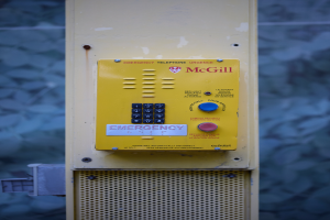 Ein gelbes Notruf-Telefon an der Seite eines Gebäudes montiert, mit Tasten und Text für Notrufe, vor einer Wand.