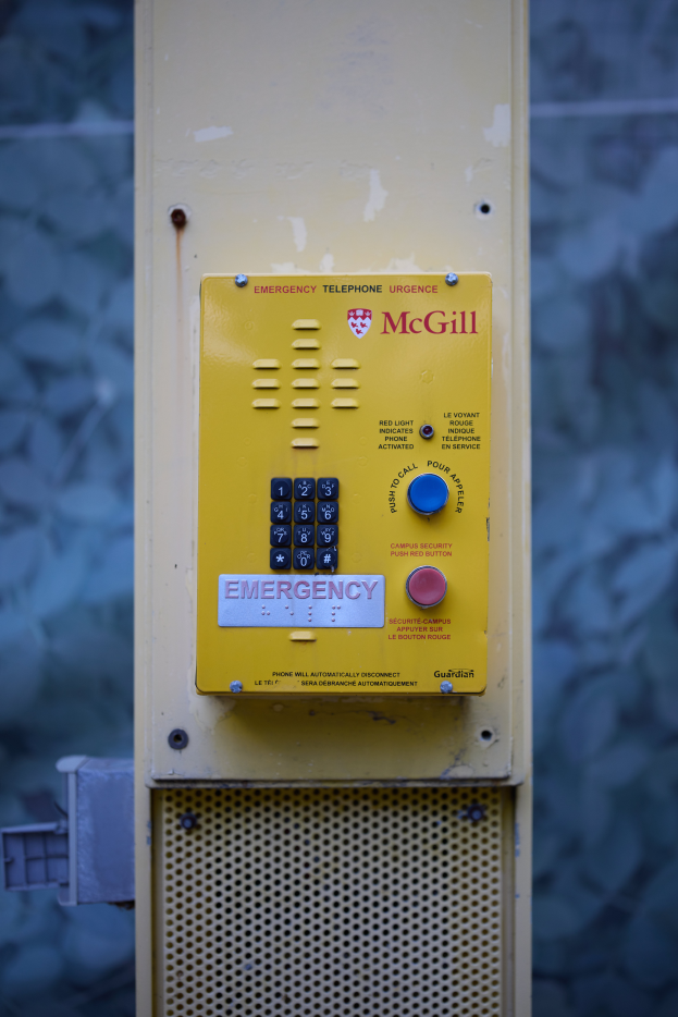 Ein gelbes Notruf-Telefon an der Seite eines Gebäudes montiert, mit Tasten und Text für Notrufe, vor einer Wand.