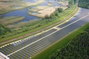 Eine Luftaufnahme eines Solarparks mit zahlreichen Solarpanelen auf einem Feld, umgeben von Bäumen, Gras und Wasser, mit einem Zug, der auf einem Bahngeleis in der Nähe fährt.