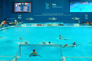 Eine Gruppe von Menschen, die Wassersport betreiben, in einem großen Pool mit einem Netz in der Mitte, Zuschauern auf der linken Seite und einer Wand mit einem Bildschirm im Hintergrund bei den 2016 FINA World Championships in Kazan.
