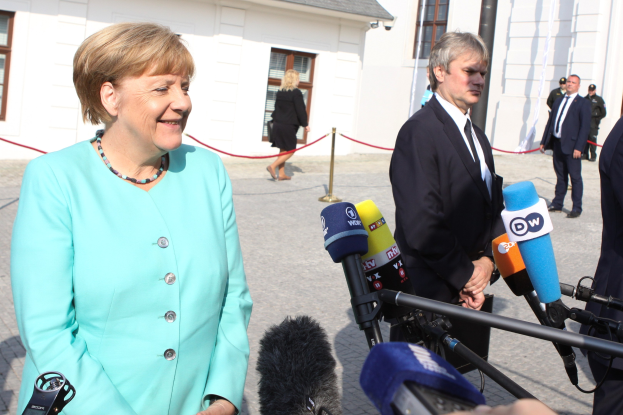 Bundeskanzlerin Angela Merkel spricht vor einem Gebäude mit einer Gruppe von Menschen, die Mikrofone halten und eine Absperrung im Hintergrund unter einem bewölkten Himmel.