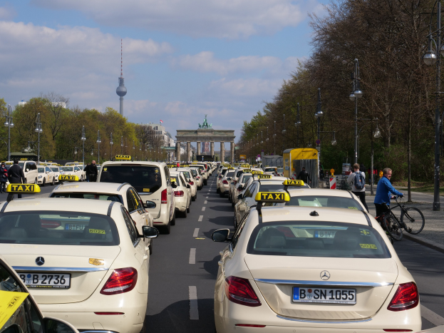 Eine lange Reihe von Taxis, die an einer belebten Straße in Berlin, Deutschland, geparkt sind, mit Fahrradfahrern und Fußgängern auf dem Gehweg, flankiert von Laternen und Bäumen, und Gebäuden, einem Bogen und einem Turm im Hintergrund.