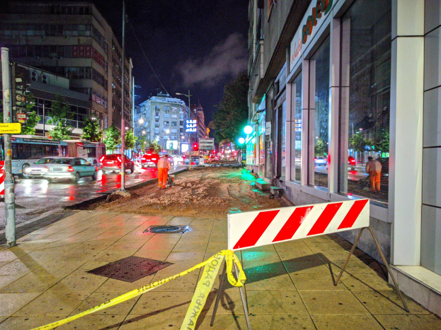 Eine nächtliche Straßenansicht einer Stadt mit gelbem Absperrband auf dem Gehweg, Fahrzeugen auf der Straße, Gebäuden mit Fenstern auf beiden Seiten, Bäumen, Laternenpfählen und Schildern mit Text, unter einem bewölkten Himmel.