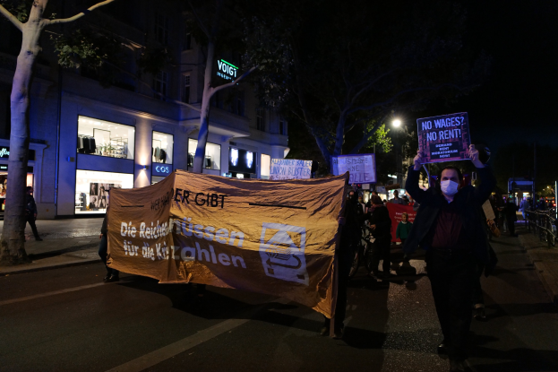 Gruppe von Menschen mit Schildern und Plakaten bei einer nächtlichen Demonstration auf einer Berliner Straße, mit Bäumen, Gebäuden, Laternen und Fahrrädern im Hintergrund.