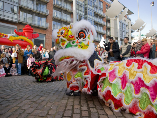 Ein lebendiges chinesisches Neujahrsfest in Amsterdam mit einer Drachentänzer-Vorführung und einer Menge Schaulustiger, einige halten Kameras, vor einem Hintergrund aus Gebäuden, Laternenmästen und einem klaren blauen Himmel.