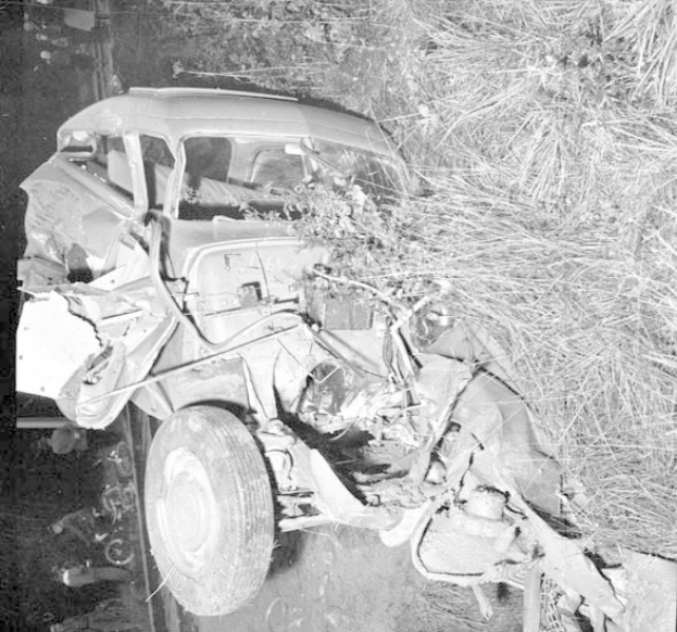 Schwarze und weiße Fotografie eines stark beschädigten Autos mit eingedellter Front und zersplitterten Fenstern am Straßenrand, mit einem Baum und verstreuten Trümmern im Hintergrund.