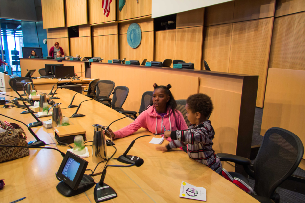 Zwei Kinder sitzen an einem langen Tisch mit Mikrofonen, Flaschen, Taschen und Papieren und führen eine Diskussion bei einer Schulratssitzung, mit Menschen und Fahnen im Hintergrund.