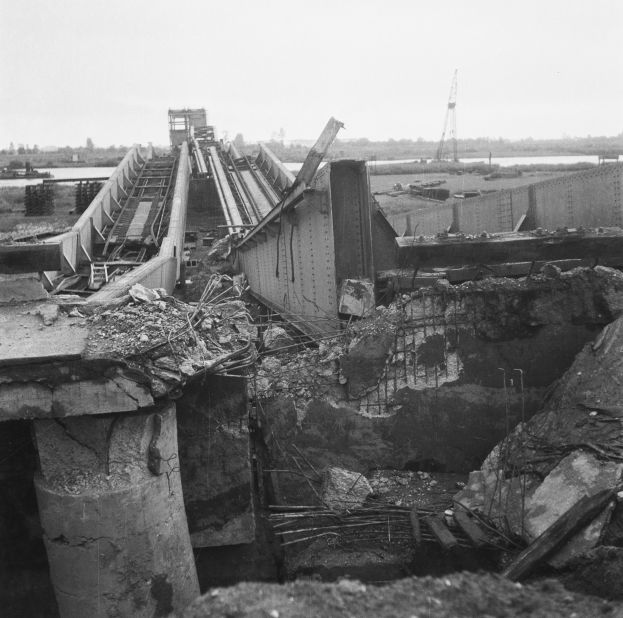 Ein Schwarz-Weiß-Foto einer teilweise gesprengten Brücke mit verstreuter Trümmer, das Wasser, Bäume, einen Turm und den Himmel im Hintergrund zeigt.