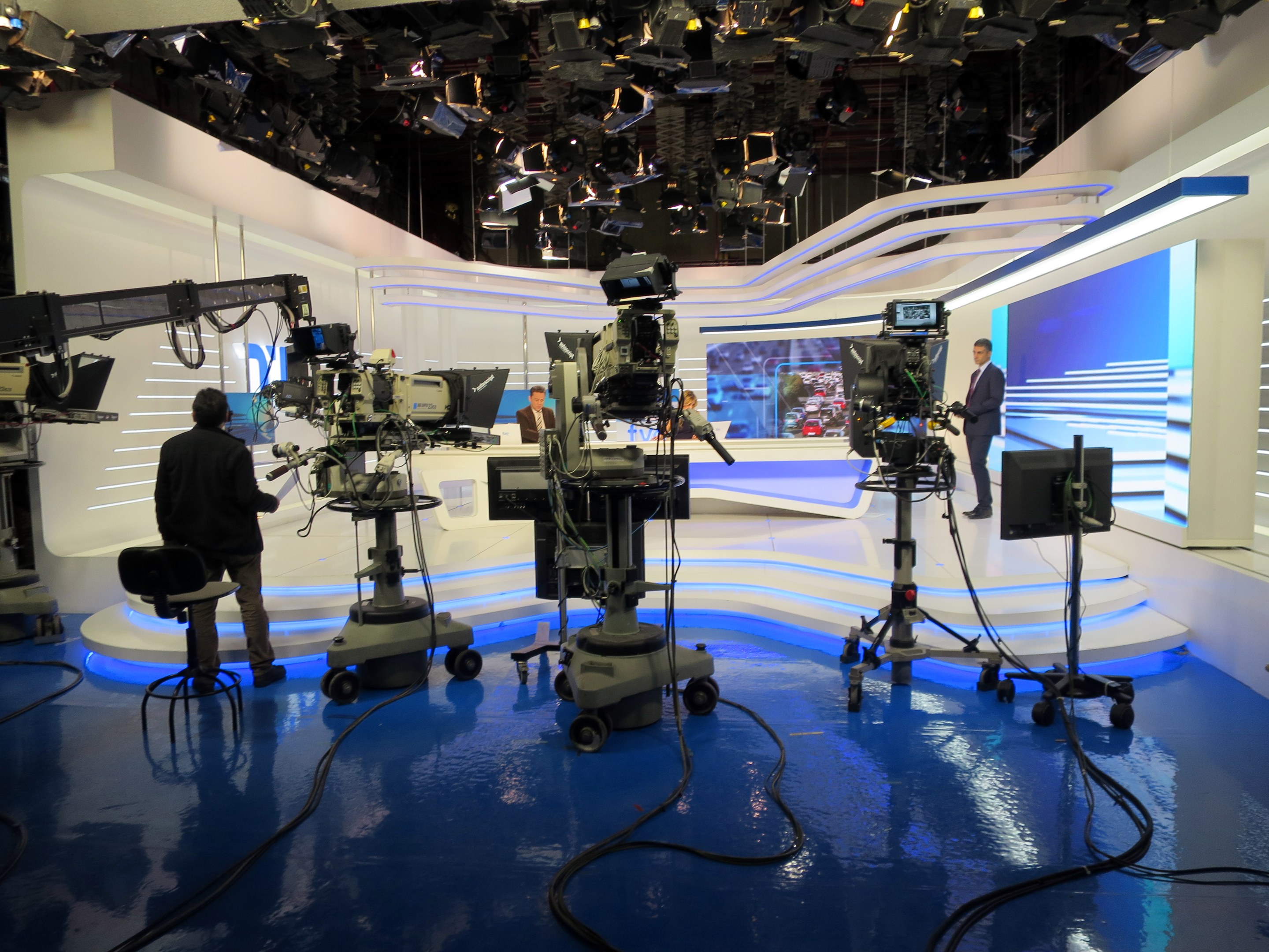 Ein TV-Studio mit mehreren Kameras auf dem Boden, einer Person im Vordergrund und mehreren Personen im Hintergrund, mit Deckenleuchten, die eine Livesendung anzeigen.