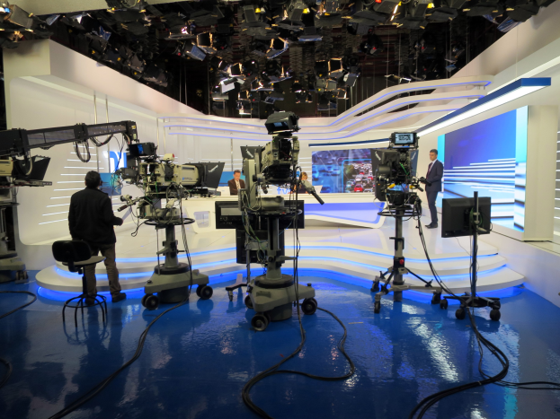 Ein TV-Studio mit mehreren Kameras auf dem Boden, einer Person im Vordergrund und mehreren Personen im Hintergrund, mit Deckenleuchten, die eine Livesendung anzeigen.