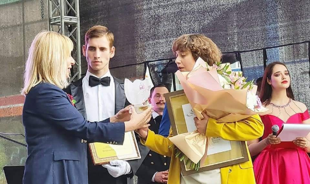 Eine Gruppe von Menschen auf einer Bühne, wobei einer in einem gelben Kleid Blumen hält und ein anderer Papiere und ein Mikrofon, was auf eine Preisverleihung hindeutet.