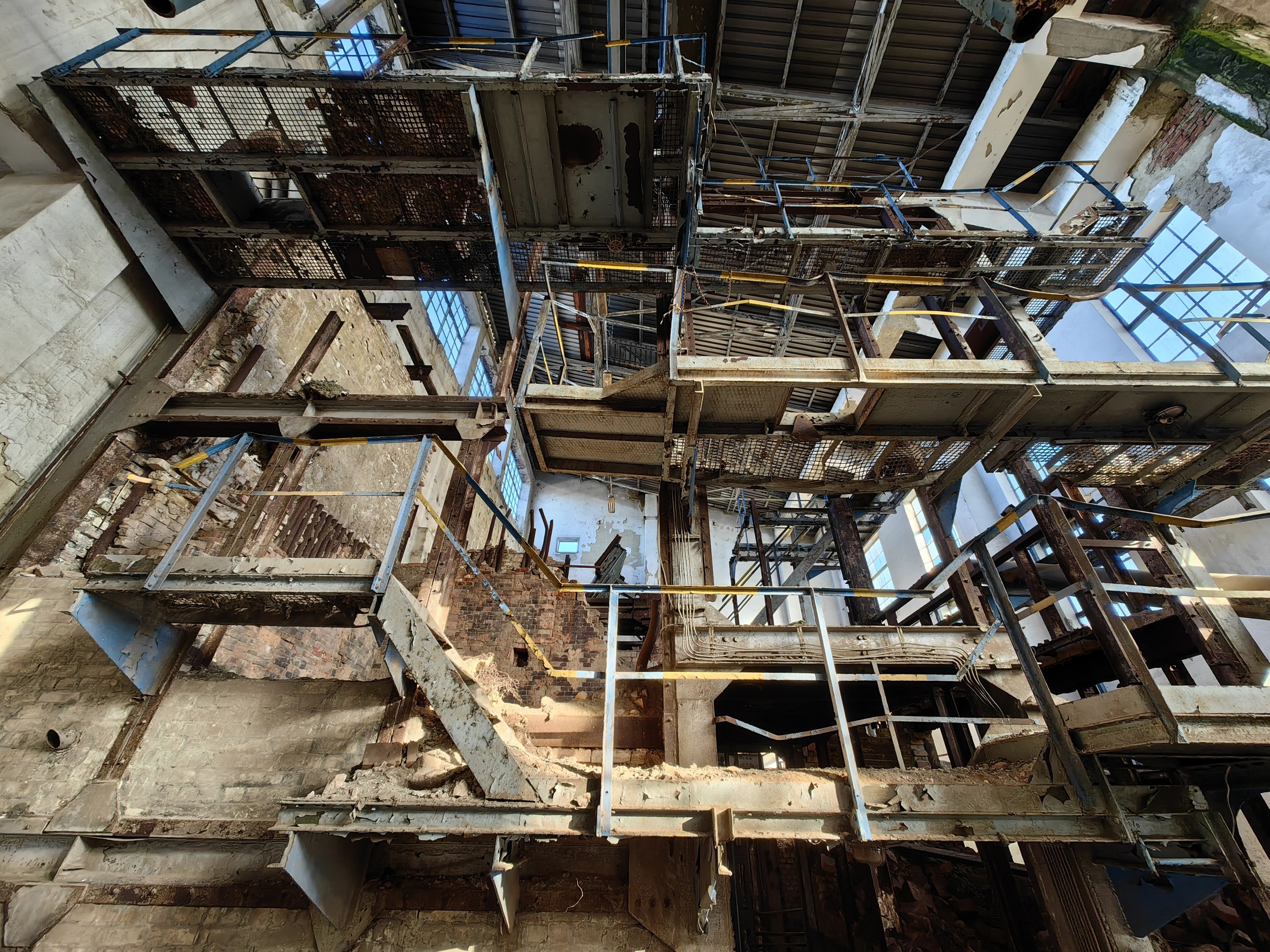 Altes Industriegebäude mit einem zentralen Metallgerüst, umgeben von verrosteten Eisenstangen, Treppen, Wänden und Fenstern, die Zeichen von Verfall und Verwahrlosung zeigen.