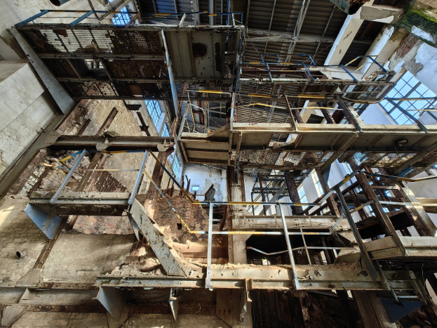 Altes Industriegebäude mit einem zentralen Metallgerüst, umgeben von verrosteten Eisenstangen, Treppen, Wänden und Fenstern, die Zeichen von Verfall und Verwahrlosung zeigen.