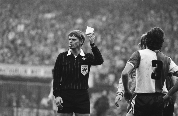 Schwarzes und weißes Bild eines Schiedsrichters mit roter Karte vor einer Menge von Menschen in Fußballuniformen mit einem Zaun im Hintergrund.