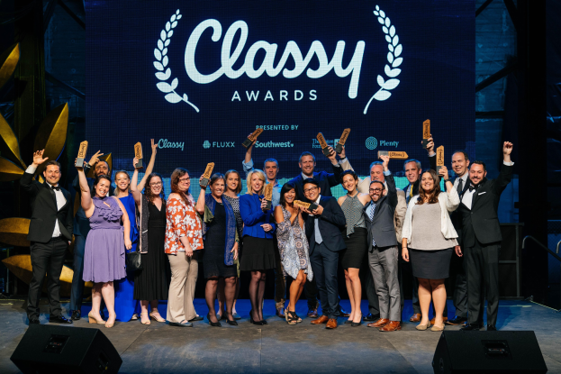 Gruppe von Menschen auf einer Bühne mit Preisen, darunter Lautsprecher und ein Bildschirm mit der Aufschrift "Classy Awards 2019" im Hintergrund.