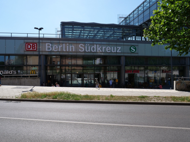 Gro├čes gl├Ąsernes Berliner Südkreuz-Bahnhof mit Text an seiner Fassade, umgeben von Laternenmasten, Lichtern, Fahrzeugen, Passanten, Fahrr├Ądern, B├Ąumen und einem klaren blauen Himmel.