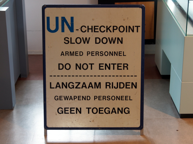 Schild auf dem Boden mit der Aufschrift "UN-Checkpoint Slow Down Armed Personnel - Do Not Enter" mit einem Glastisch und Gegenständen rechts und einer Wand mit einem Gegenstand im Hintergrund.