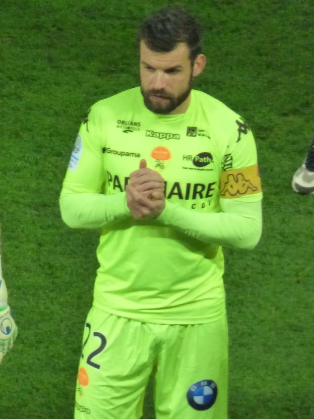 Ein Torwart in einem gelben Trikot klatscht auf einem Fußballfeld, trägt ein grünes T-Shirt und Shorts und hat ein weiteres Bein links im Bild.