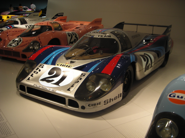 Ein Porsche 917K Le Mans Rennauto wird in einem Museum ausgestellt, umgeben von anderen Sportwagen und Menschen, mit einer Wand und einem Papier mit Text im Hintergrund.