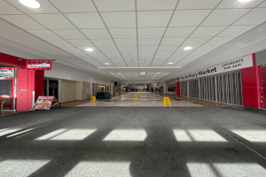 Innenansicht eines hell erleuchteten Einkaufszentrums mit einem Stuhl und einer Tafel "Sydbury Market - Vorher und Nachher" links und verstreuten Gegenständen rechts und auf dem Boden.