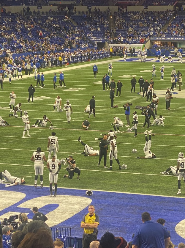 Ein Fußballspiel in einem Stadion mit Spielern auf dem Feld und Zuschauern im Hintergrund, einschließlich eines Kameramannes, der die Live-Übertragung anzeigt.