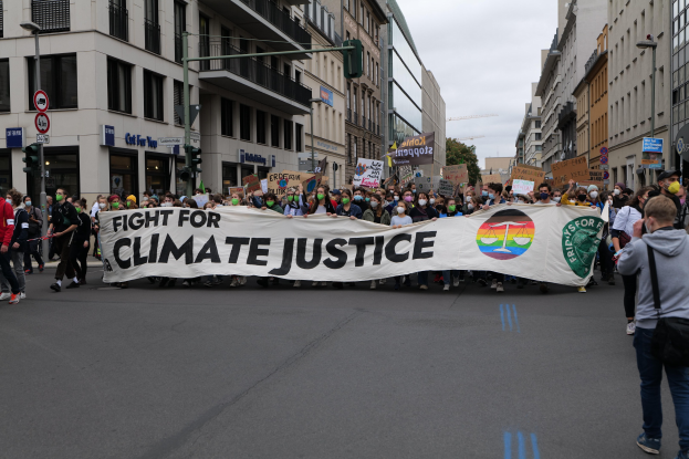 Eine Gruppe von Menschen marschiert auf einer Straße und hält ein "Kampf für Klimagerechtigkeit"-Schild, mit Gebäuden, Ampeln, Bäumen und einem bewölkten Himmel im Hintergrund.